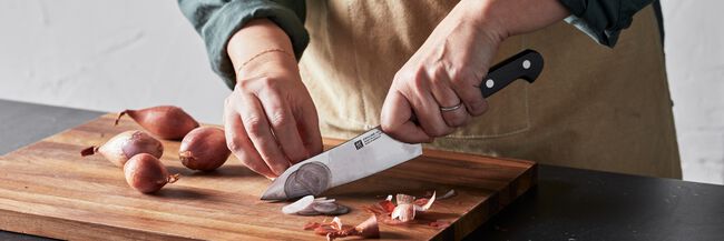 NEW ZWILLING Pro 5.5 in Rocking Santoku high-quality German-made Knife slicing shallots on a cutting board. On Sale now!
